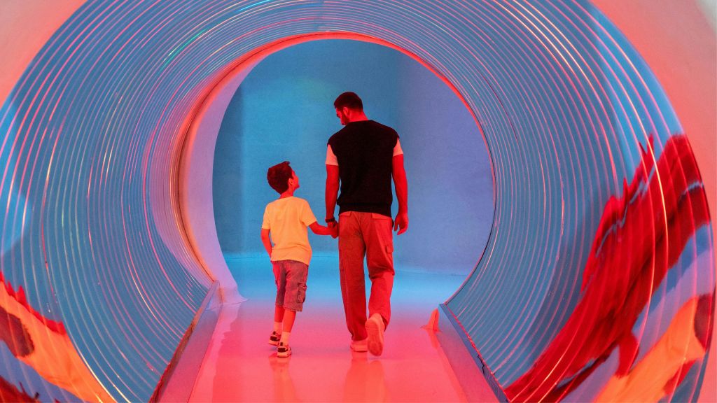 어른과 어린이가 손을 잡고 휘어진 벽과 반사 바닥이 있는 밝고 화려한 터널을 걷고 있습니다.