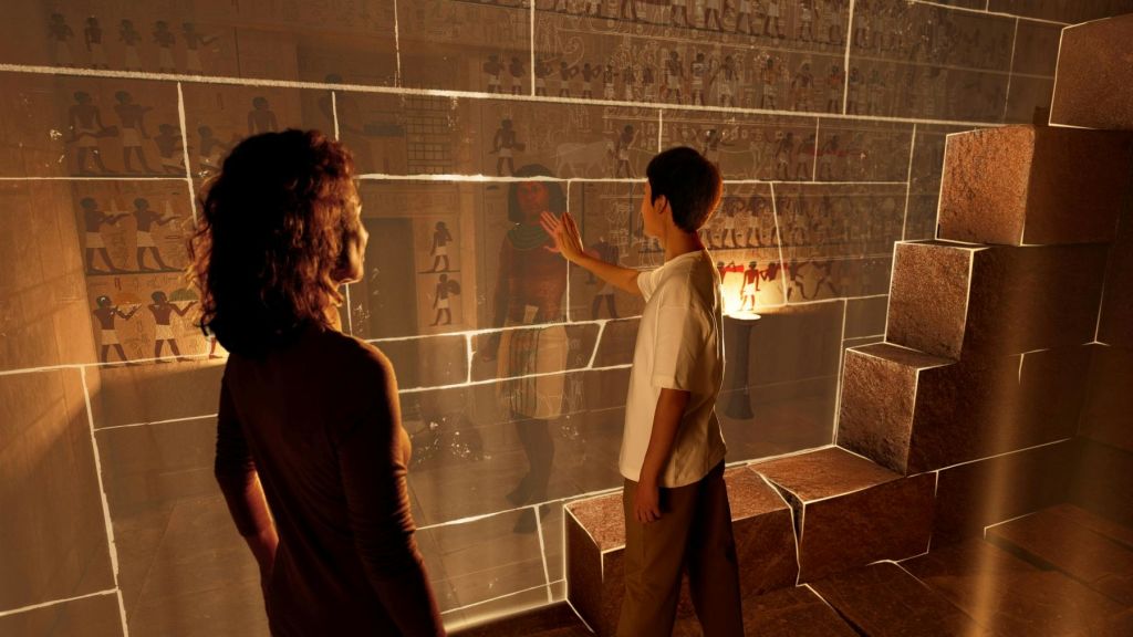 한 남성이 고대 이집트 그림을 전시하는 유리 벽을 만지는 동안 한 여성이 지켜보고 있습니다. 배경에는 스톤 블록과 램프가 있습니다.