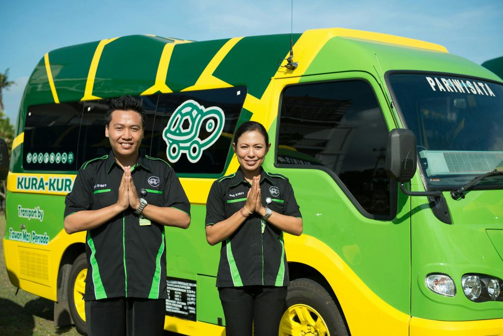 유니폼을 입은 두 사람이 거북이 로고가 있는 녹색과 노란색 차량 앞에서 나마스테 제스처로 손을 잡고 서 있습니다.