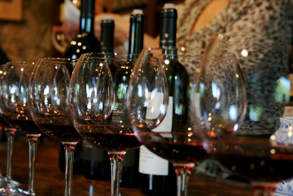 나무 표면에 와인병 몇 개 앞에 레드 와인을 가득 담은 와인잔 네 개를 배경으로 흐릿한 인물이 등장합니다.