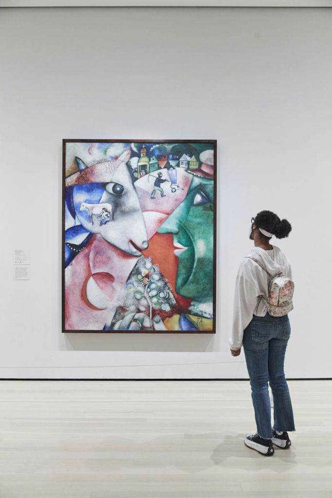 흰색 후드티와 배낭을 착용한 채 미술관에서 다채로운 추상화를 보고 있는 사람.