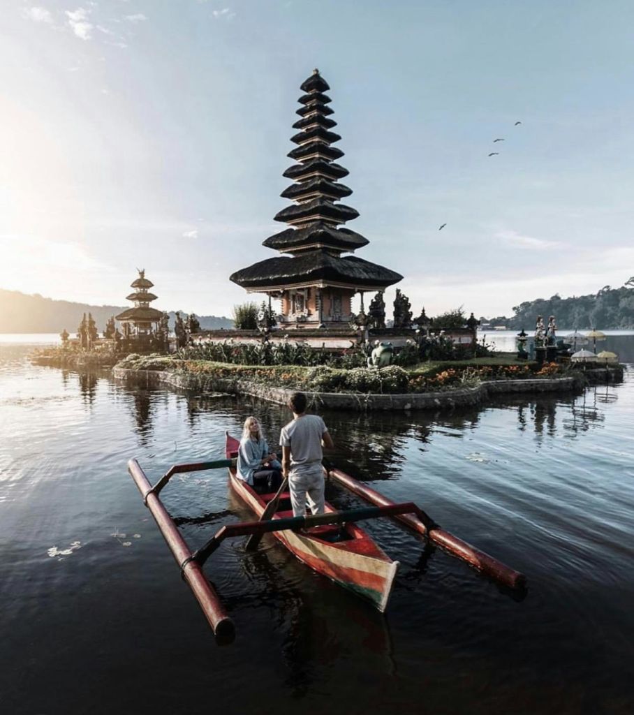 산과 하늘을 배경으로 고요한 호수의 발리 사원에 근접한 작은 보트를 탄 두 사람.