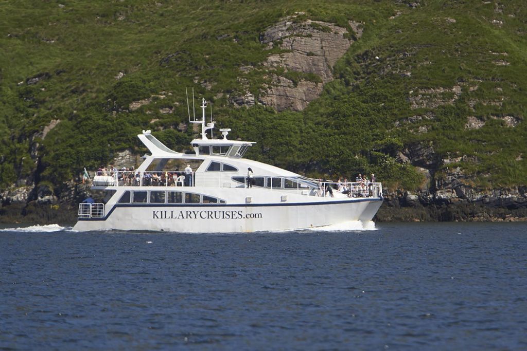 바위가 많고 푸른 언덕 근처의 갑판 돛에 승객이 타고 있는 흰색 킬러리 크루즈 보트.