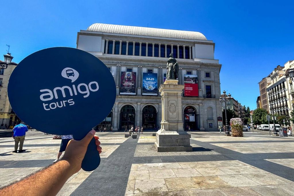 햇살이 내리쬐는 광장에 있는 동상이 있는 유서 깊은 건물 앞에서 '아미고 투어' 라는 표지판을 들고 있는 손.