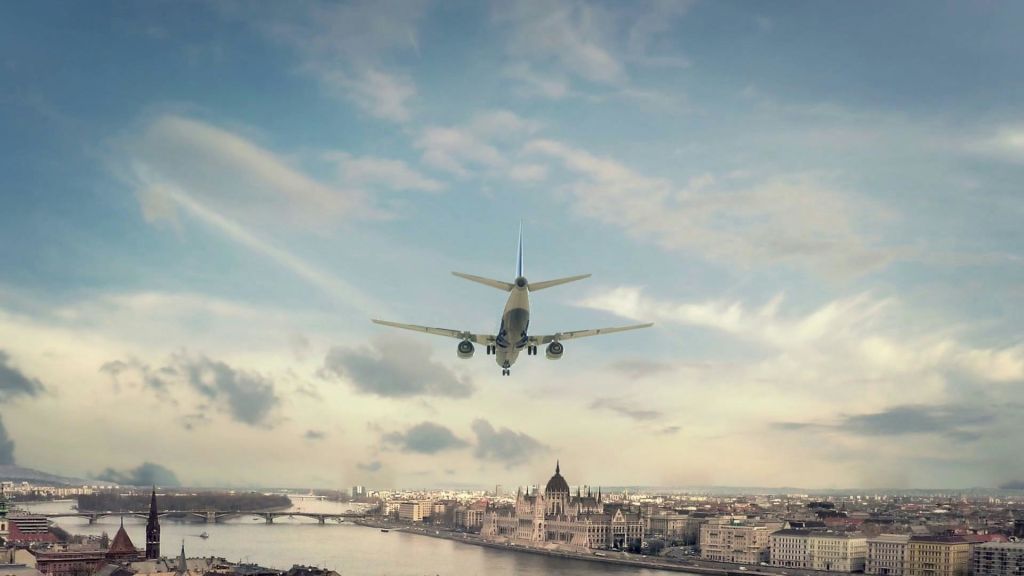 강과 건물, 부분적으로 흐린 하늘이 있는 도시 풍경 위로 낮게 날아가는 비행기.