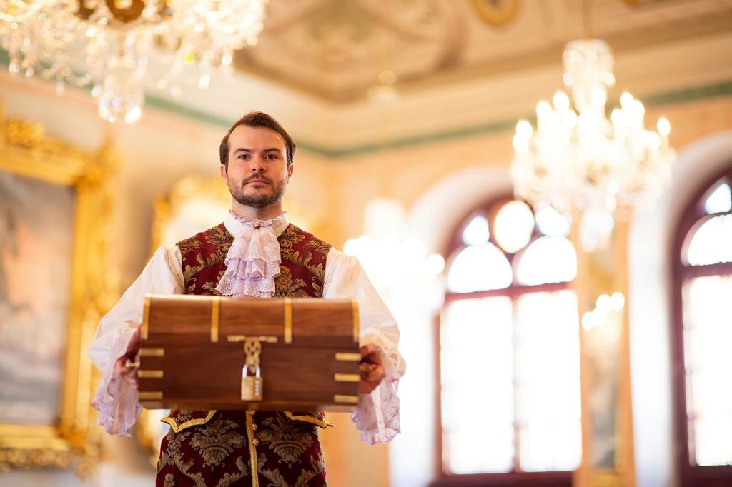 시대 의상을 입은 남자는 샹들리에와 액자에 담긴 예술품이 있는 화려한 방에서 나무 상자를 들고 있습니다.