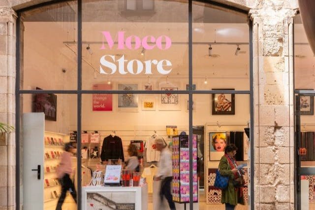 "Moco Store" 가 전시된 유리 외관이 있는 상점가에는 예술품과 상품이 전시되어 있습니다. 한 사람이 바깥을 지나갑니다.