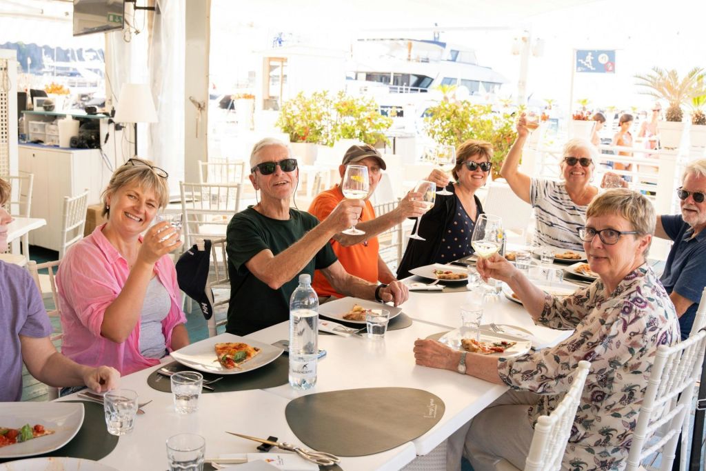 6명의 일행이 야외 테이블에 둘러앉아 경쾌한 토스트에 와인잔을 올리고 식사를 즐깁니다.