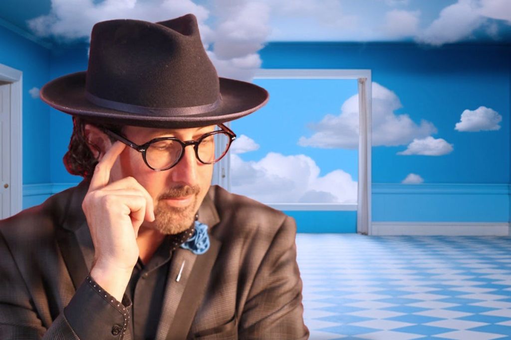파란 벽과 구름이 그려진 하늘 벽화가 있는 방에서 안경과 검은 모자를 쓴 남성.