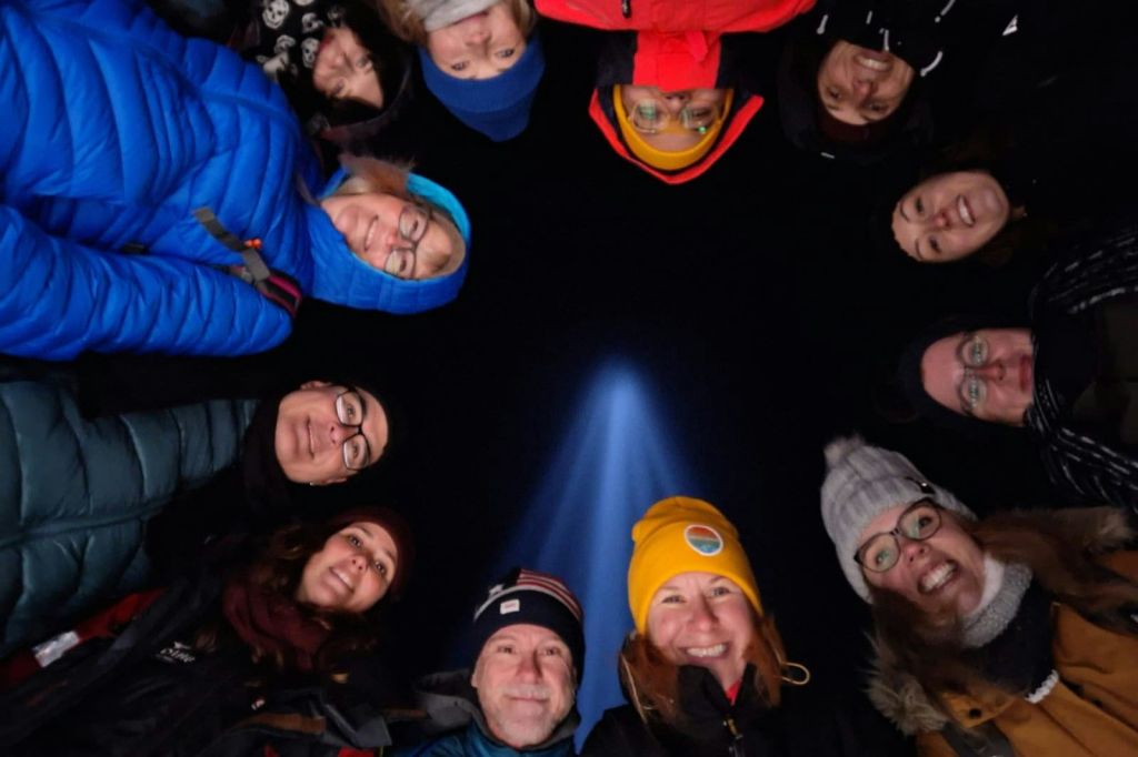 한 무리의 사람들이 원을 그리며 카메라를 향해 미소를 짓고 있습니다. 배경에는 이매진 피스 타워 (Imagine Peace Tower) 의 빛이 있습니다.