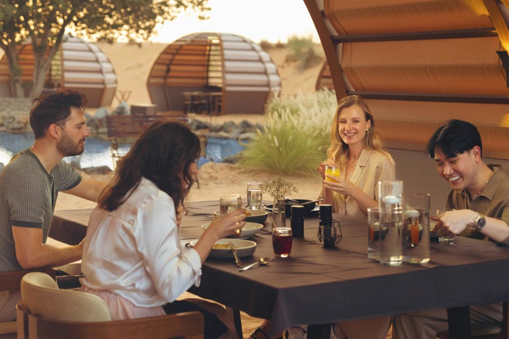 돔형 구조물을 배경으로 사막을 배경으로 야외 테이블에서 식사를 즐기는 세 사람.