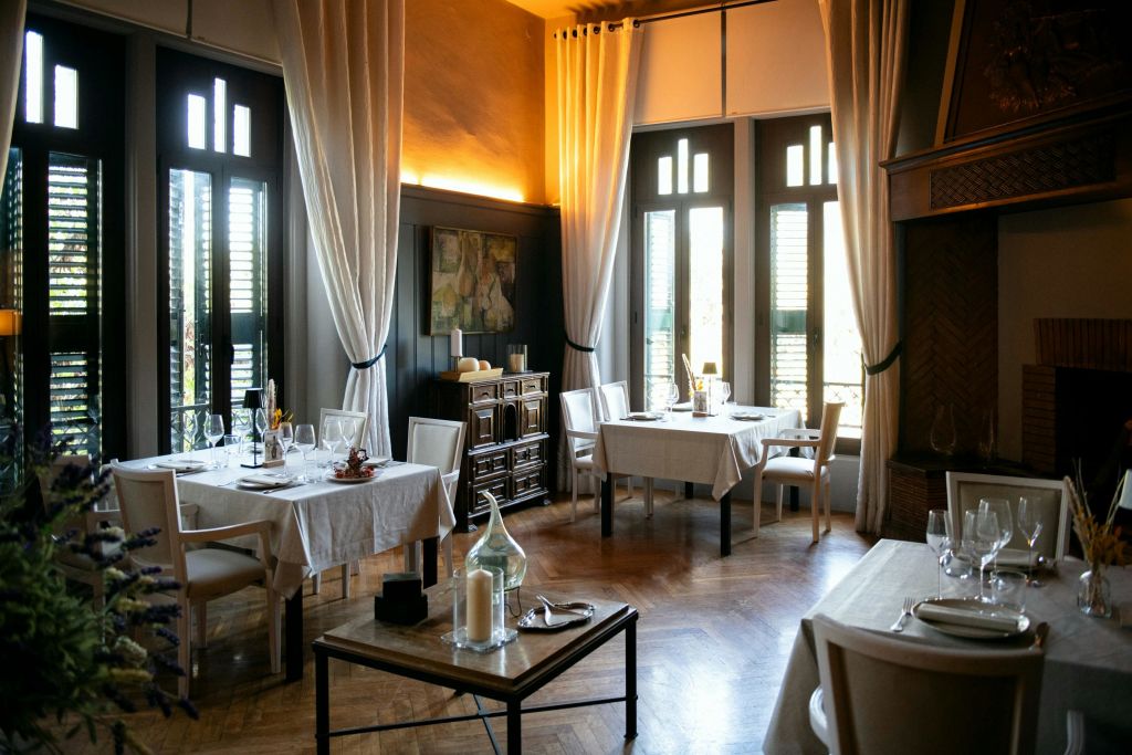 우아한 흰색 식탁보, 목재 가구, 높은 창문으로 들어오는 부드러운 자연 채광이 특징인 아늑한 레스토랑 인테리어.