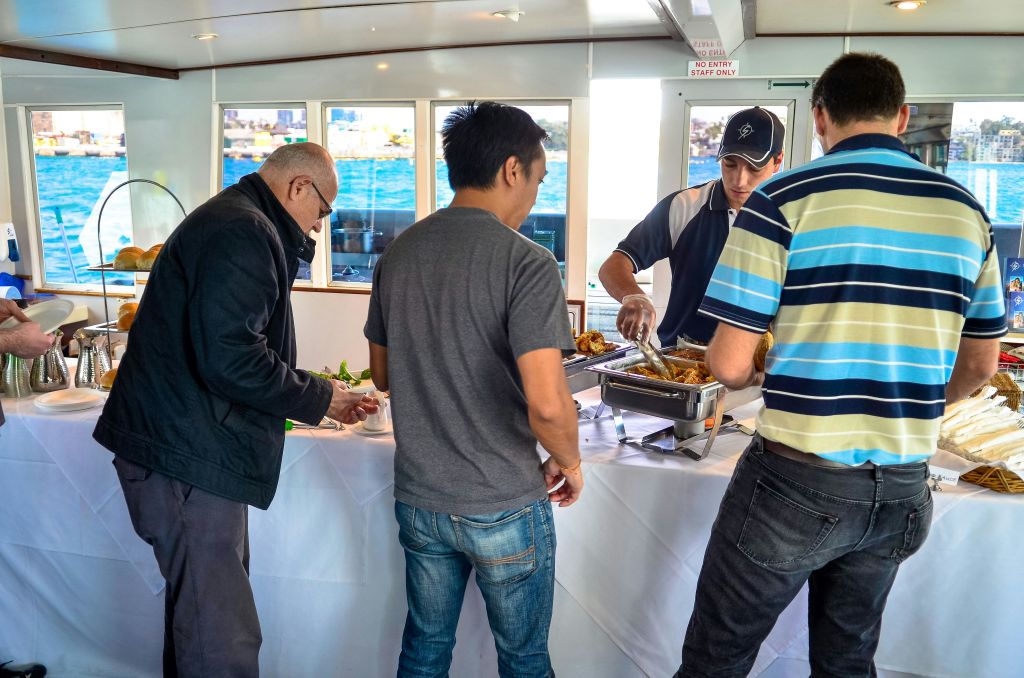 세 명의 남성이 뷔페 테이블에 앉고, 한 명은 음식을 대접하고, 다른 한 명은 기다립니다. 창문이 있는 보트에 세워진 풍경은 바깥의 물을 보여줍니다.