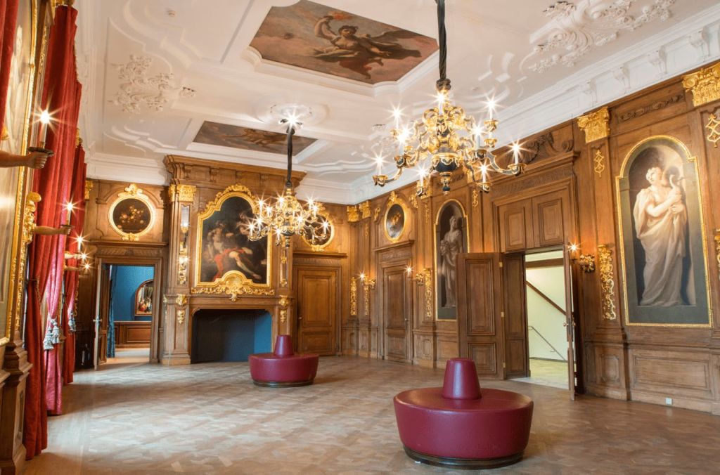 샹들리에, 벽과 천장에 그려진 그림, 조각된 나무 벽, 둥근 빨간색 좌석 공간이 있는 화려한 객실입니다.