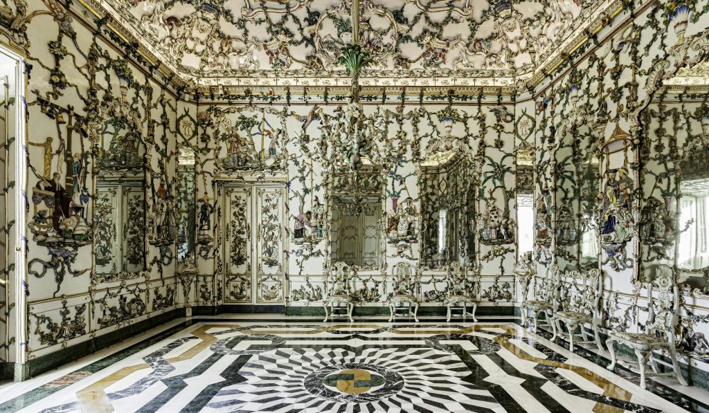 정교한 벽과 천장 디자인, 거울, 샹들리에, 흰색 의자, 흑백 패턴의 바닥이 있는 화려한 객실입니다.