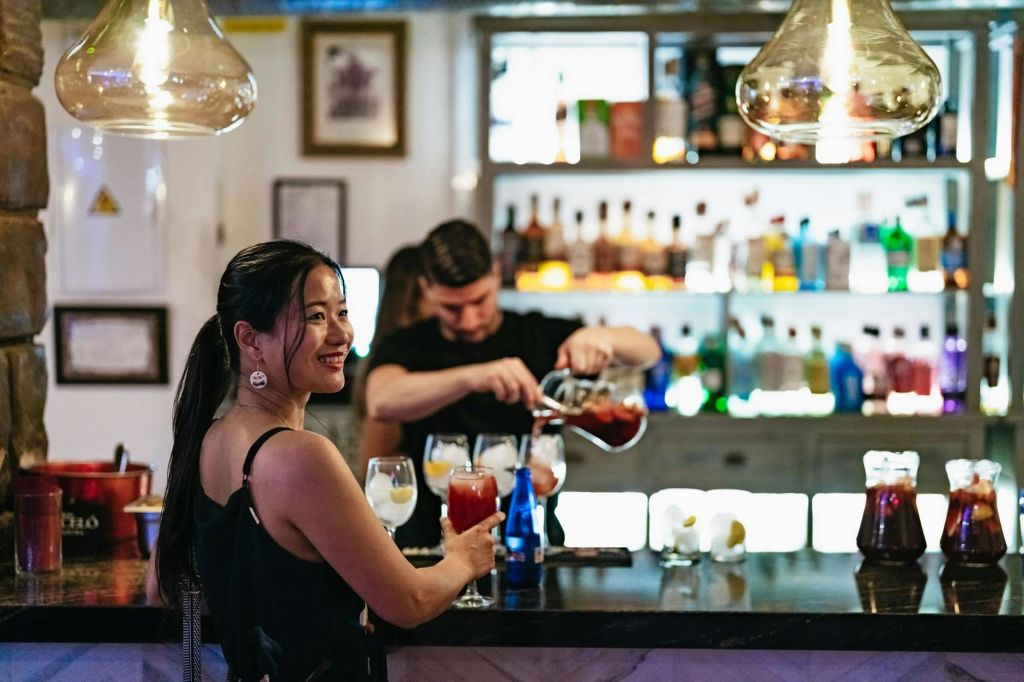 한 여성이 바에서 미소를 짓고 있고, 바텐더가 셰이커를 배경으로 음료수를 붓고 있습니다. 그 뒤로 병 선반이 보입니다.