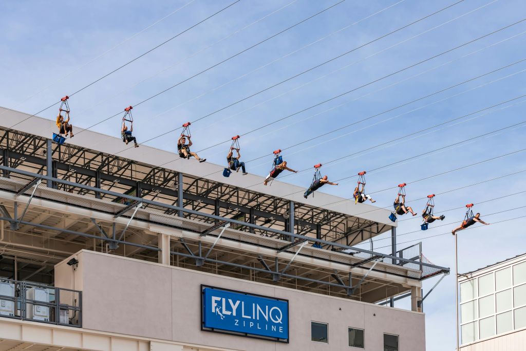 "Fly LINQ Zipline" 이라는 표지판과 배경에 맑고 푸른 하늘이 있는 건물 위에서 사람들이 집핑을 하고 있습니다.