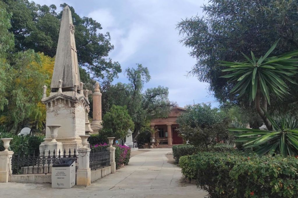 나무, 수풀, 기념물로 둘러싸인 돌 통로가 있는 야외 공원 풍경. 테라코타 건물이 배경으로 보입니다.