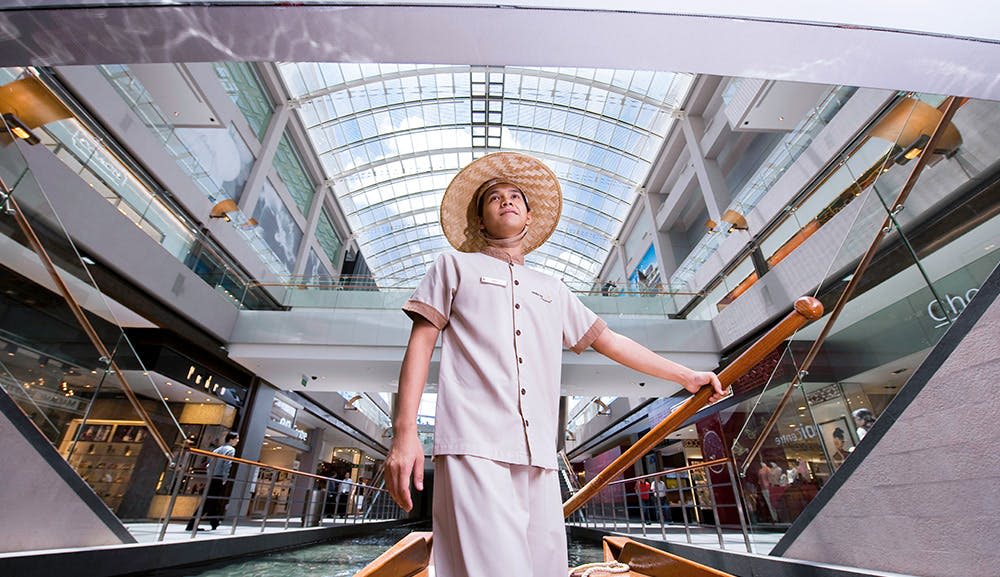 베이지색 유니폼과 밀짚 모자를 쓴 남성이 현대적인 유리 지붕 쇼핑몰 내부의 보트에 서 있습니다.