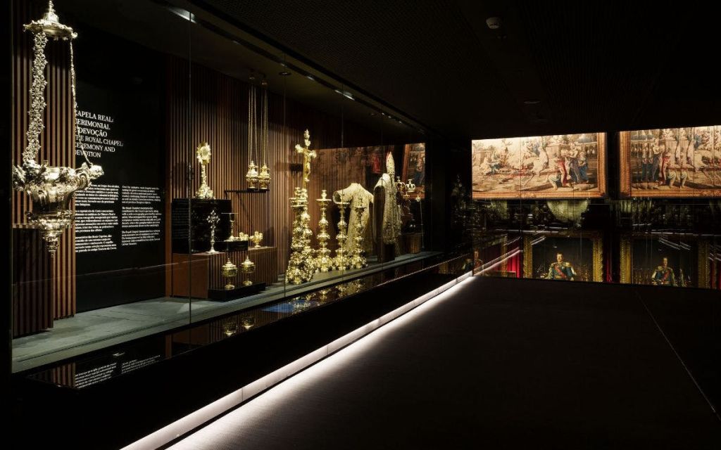 태피스트리를 배경으로 화려한 황금 종교 유물과 가운이 전시된 조명이 희미한 박물관입니다.