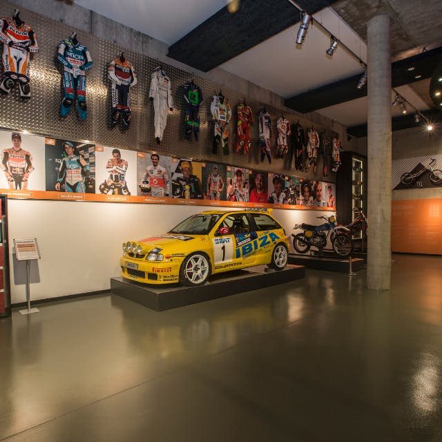 빈티지 레이싱 장비, 노란색 랠리 차량, 벽에 걸린 레이싱 슈트, 플랫폼에 달린 오토바이를 전시하는 박물관 전시물입니다.