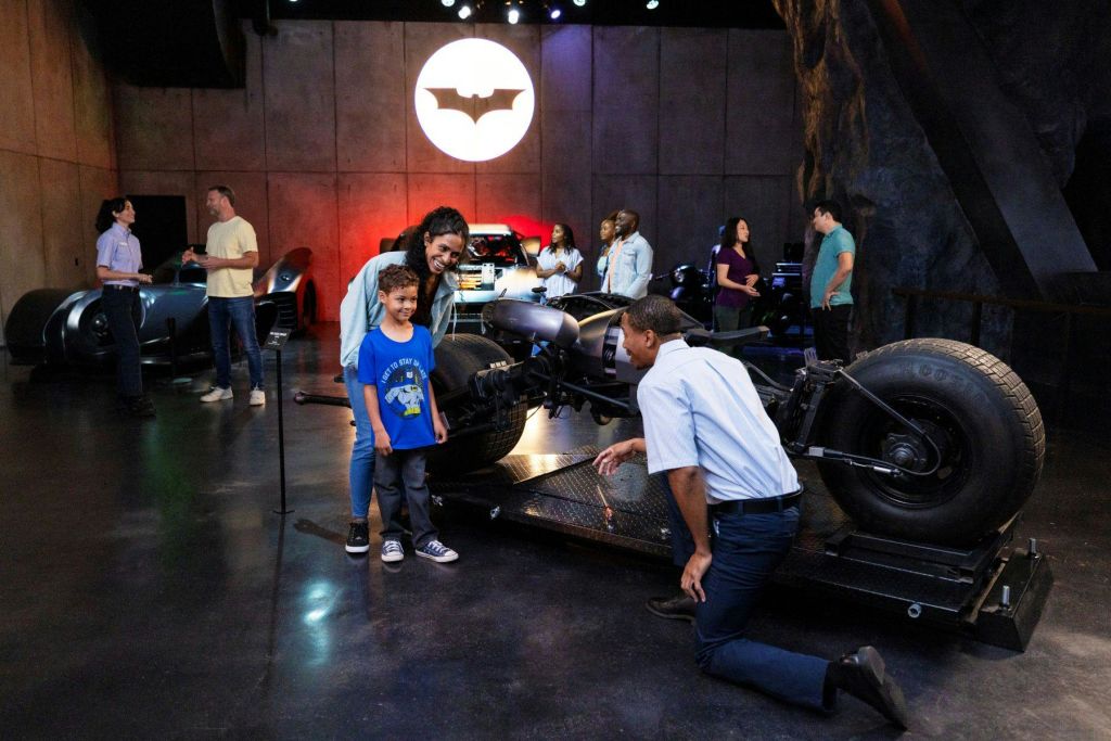 벽에 커다란 박쥐 신호가 있는 배트맨 테마 전시회에서 차량과 디스플레이를 보고 상호작용하는 사람들.