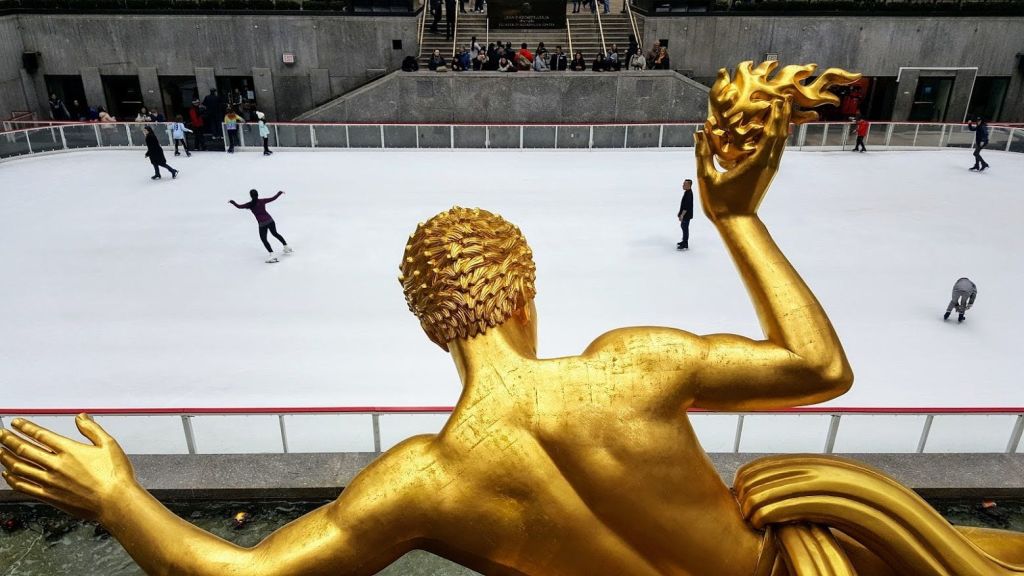 빙상 링크가 내려다보이는 전경의 황금 조각상에는 스케이트를 타는 사람들과 관객들이 앉아 있습니다.