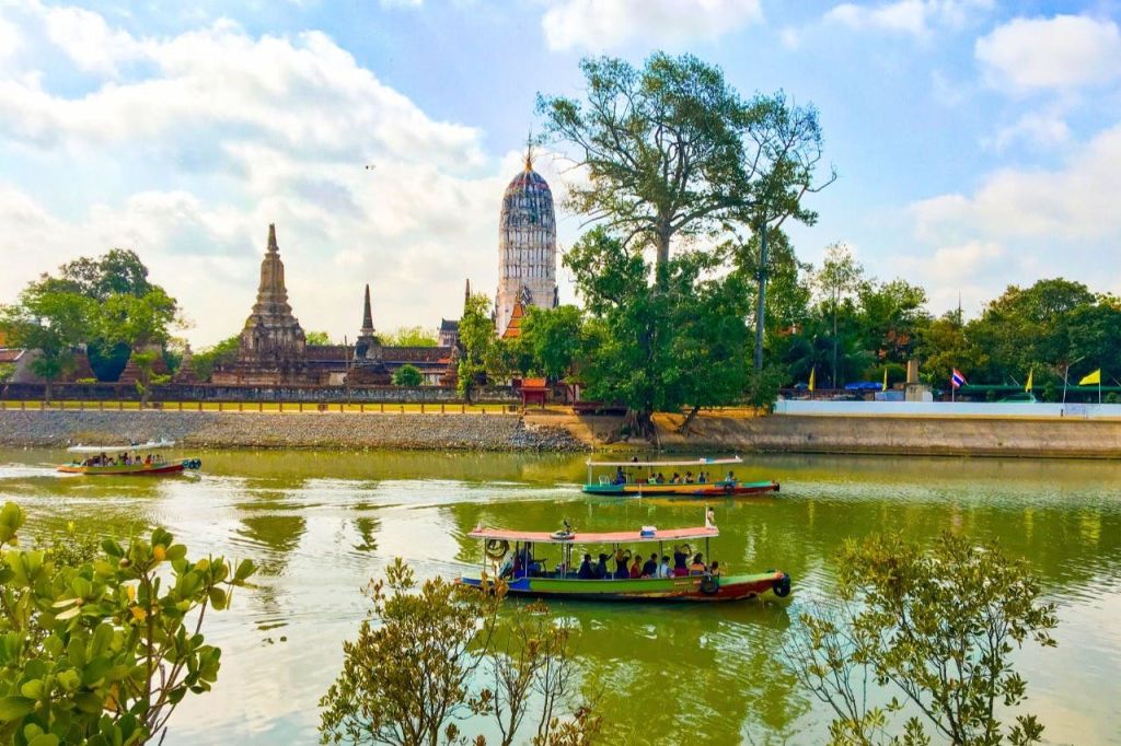 화창한 하늘 아래 고대 사원 구조물과 나무를 배경으로 강에서 승객과 함께 보트.