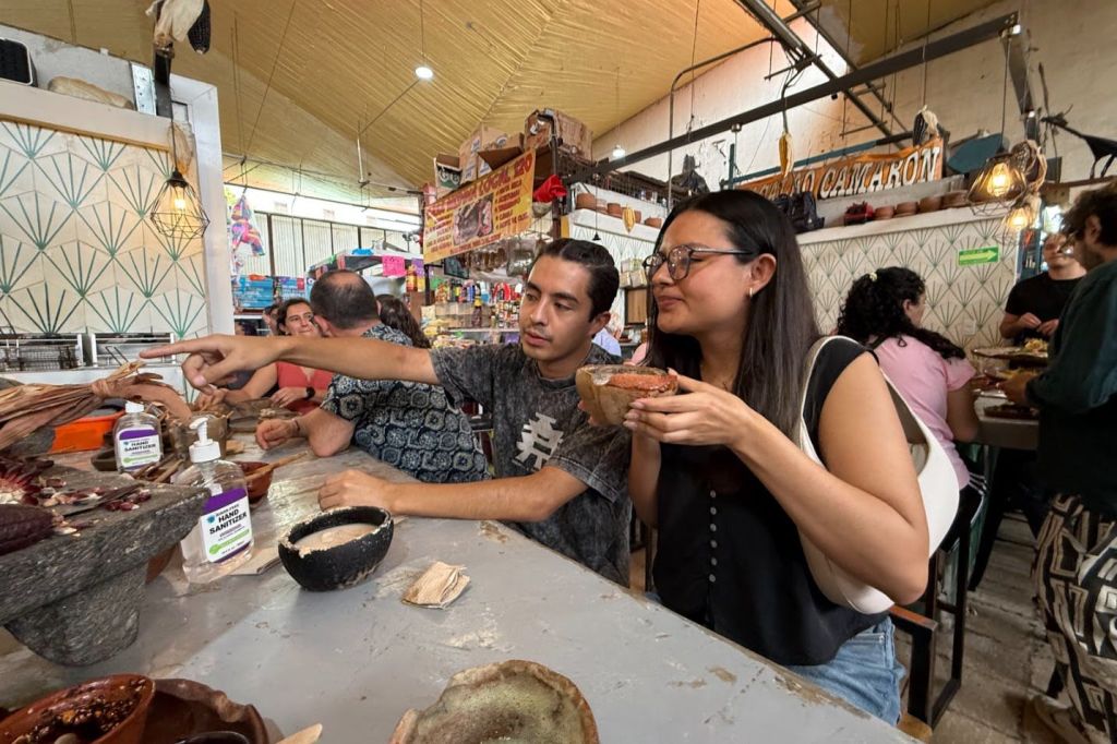 음식 가판대에 앉아 전통 그릇을 들고 있는 남성과 여성으로, 다른 후원자와 음식이 배경으로 보입니다.