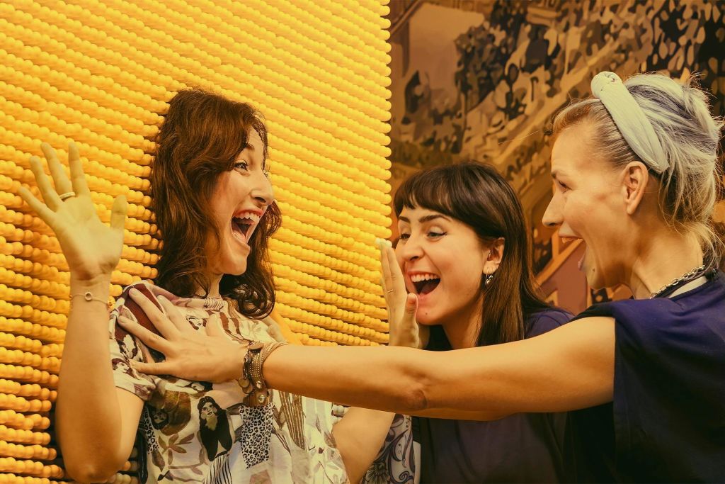 노란색 질감의 배경을 배경으로 실내에서 세 명의 여성이 신나게 인사하고 있습니다.