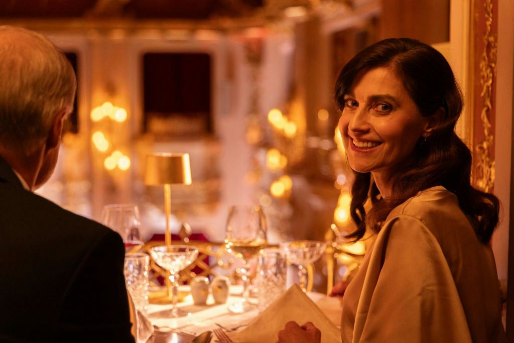 우아한 분위기의 한 여성이 카메라를 보며 미소를 짓고 있습니다. 그녀는 와인잔과 따뜻하고 분위기 있는 조명이 있는 테이블에 앉아 있습니다.