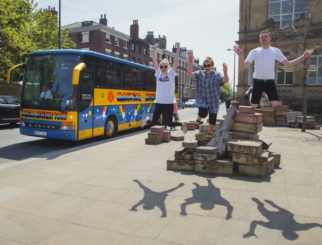 알록달록한 버스와 벽돌 더미 옆에서 세 사람이 즐겁게 뛰어놀고 있습니다. 바닥에 그림자가 보입니다.