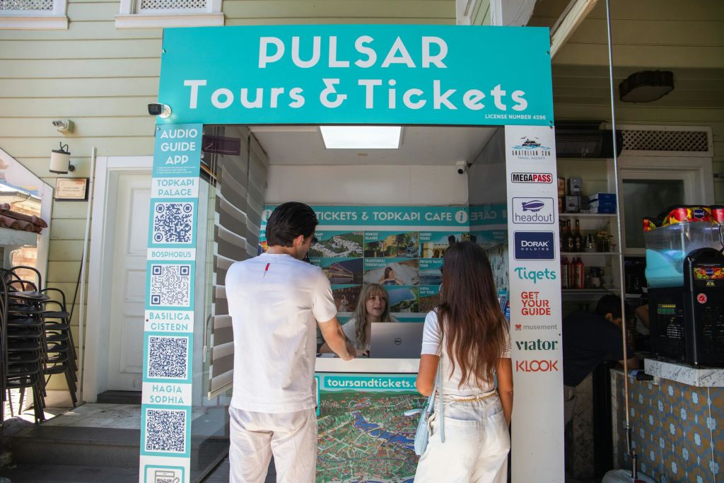 한 남녀가 '펄서 투어 & 티켓' 부스에 서서 직원과 이야기를 나누고 있습니다.