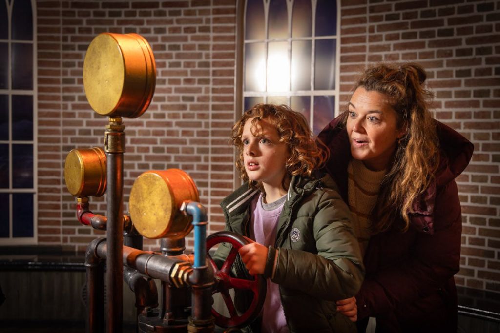 벽돌 벽과 아치형 창문이 있는 방에서 빨간 바퀴가 달린 기계 장치와 상호 작용하는 어린이와 어른.