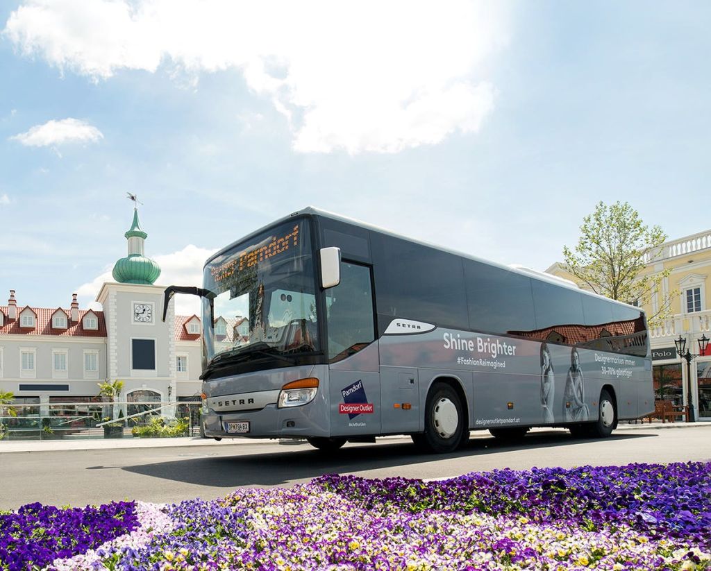 보라색 꽃 침대에 둘러싸인 시계탑이 있는 건물 근처에 '샤인 브라이터' 브랜드의 버스가 주차되어 있습니다.