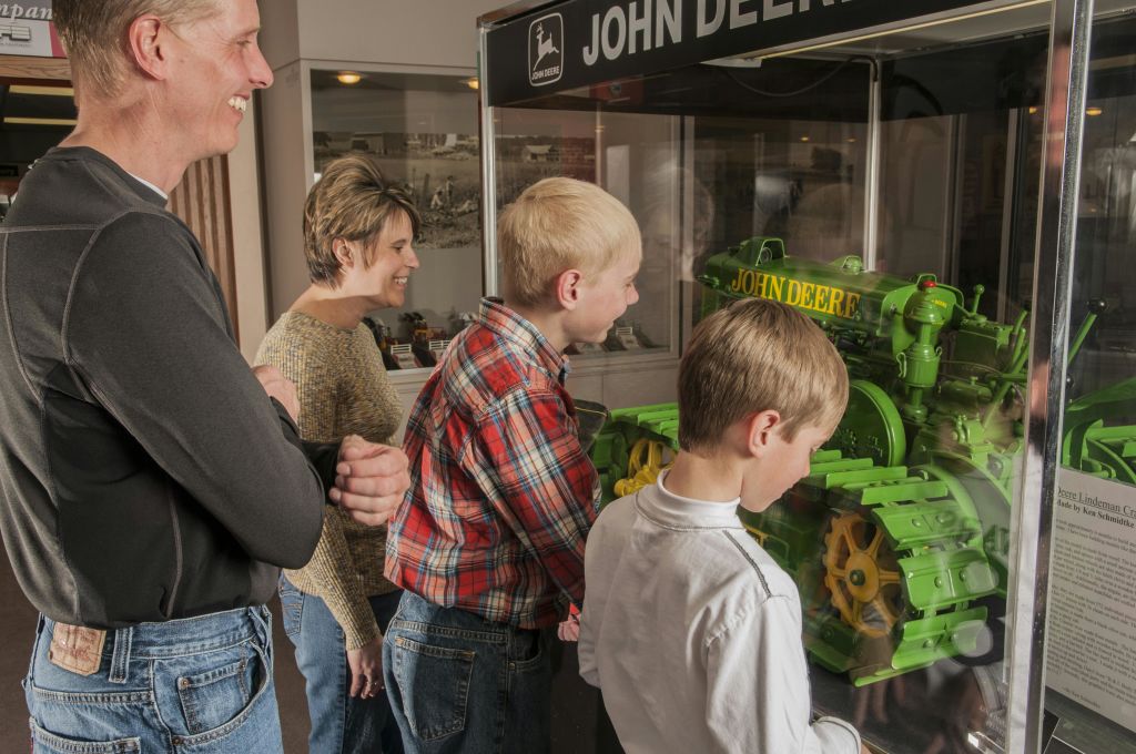 네 명의 가족이 John Deere 디스플레이를 관찰하고 두 명의 소년이 녹색 및 노란색 모델 트랙터를 면밀히 검사합니다.