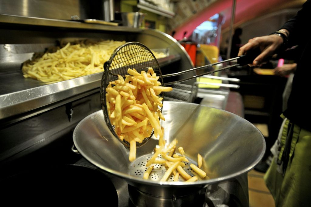 갓 조리한 감자튀김을 스트레이너가 달린 튀김기에서 금속 그릇으로 들어 올리고 감자튀김을 배경으로 더 많은 감자튀김이 나옵니다.