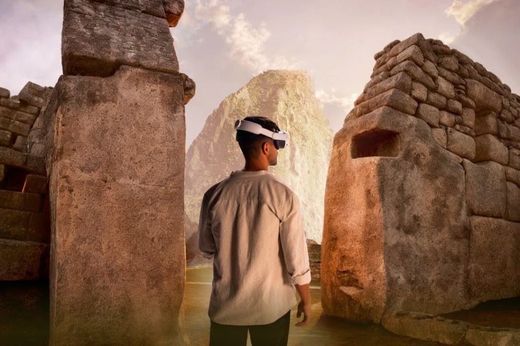 가상 현실 고글을 착용한 사람이 두 개의 커다란 석조 구조물 사이에 서 있으며, 산봉우리가 배경으로 보입니다.