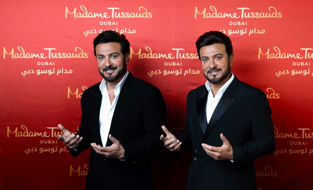 검은 양복을 입은 남성이 "마담 투소 두바이 (Madame Tussauds Dubai)" 가 반복적으로 쓰여진 빨간색 배경 앞에서 포즈를 취하고 있습니다.