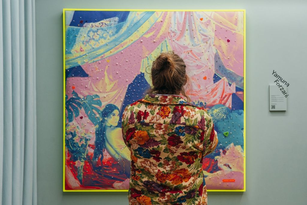 분홍색, 파란색, 노란색이 선명하게 어우러진 화려한 추상화 앞에 꽃무늬 셔츠를 입은 사람이 서 있습니다.